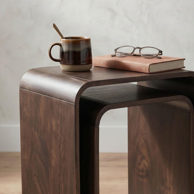 Op de afbeelding: Een donkerbruine houten bijzettafel met een gebogen ontwerp. Een keramische mok met een lepel, een boek en een bril liggen op de tafel. De mok heeft bruine en bruinachtige tinten. Het boek is lichtbruin.