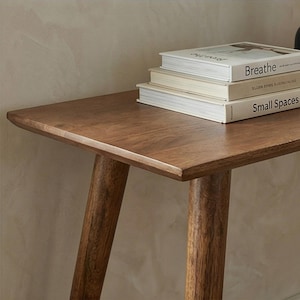 K&ouml;nnte beinhalten: Ein Beistelltisch aus Holz mit warmbraunem Finish. Der Tisch hat eine rechteckige Platte und abgewinkelte Beine. Drei B&uuml;cher sind auf dem Tisch gestapelt, das oberste Buch tr&auml;gt die Aufschrift "Breathe" und das unterste Buch "Small Spaces".