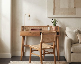 May include: A wooden desk with a cane back panel and a single drawer, paired with a matching wooden chair featuring a woven cane seat and backrest. A brass lamp, two stacked books, and a small potted plant sit on the desk surface.