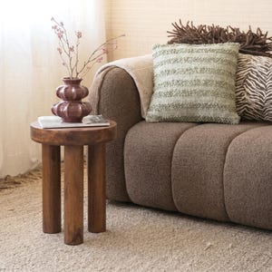 May include: A small, round wooden side table with three legs supports a plum-coloured vase holding delicate pink branches. A magazine rests on the table. A brown sofa with decorative cushions is in the background, creating a cosy living room scene.