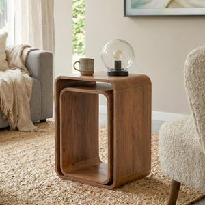 Könnte beinhalten: Ein Satz von zwei ineinandergreifenden Tischen aus warm getöntem Holz. Die Tische haben ein abgerundetes, modernes Design. Eine klare Glaskugellampe mit schwarzem Sockel und eine Keramiktasse stehen auf dem oberen Tisch.
