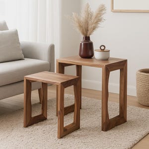May include: Two wooden side tables of different sizes. The larger table holds a dark red vase with dried ornamental grass and a small white container with a wooden lid. The tables are made of a warm-toned wood.