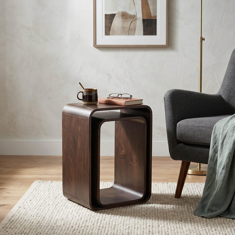 Op de afbeelding: Een donkerbruine, moderne bijzettafel met een gebogen ontwerp. Een boek en een bril liggen bovenop, samen met een mok met een lepel. De tafel staat op een cr&egrave;mekleurig tapijt, met een grijze fauteuil en kunstwerk op de achtergrond.