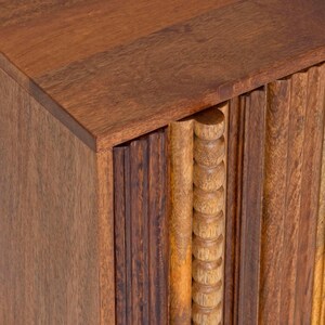 Pu&ograve; includere: Primo piano di un mobile in legno con una finitura marrone calda e ricca. Il mobile presenta una combinazione di dettagli scolpiti verticali e arrotondati, creando un design strutturato e visivamente accattivante. I pannelli superiori e laterali sono lisci.