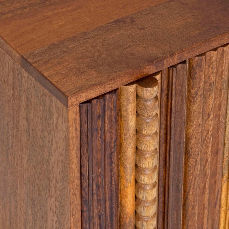 Pu&ograve; includere: Primo piano di un mobile in legno con una finitura marrone calda e ricca. Il mobile presenta una combinazione di dettagli scolpiti verticali e arrotondati, creando un design strutturato e visivamente accattivante. I pannelli superiori e laterali sono lisci.