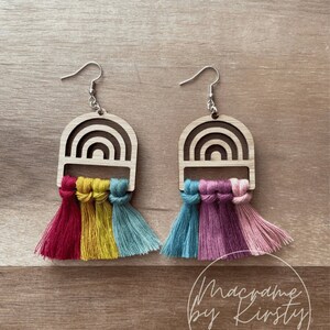 May include: A pair of rainbow-coloured wooden earrings with macrame tassels. The earrings are shaped like arches and have a woodgrain finish. The tassels are made of red, yellow, blue, pink, and teal yarn.