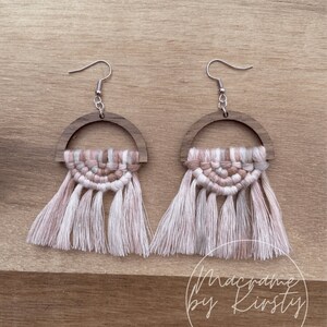 May include: A pair of pink and white macrame earrings with wooden half-circle accents. The earrings are handmade and feature a fringe design.
