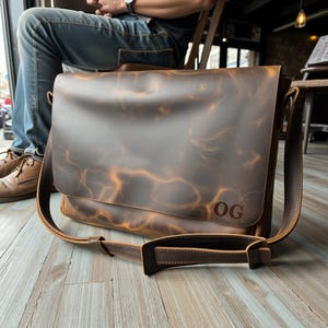 May include: A brown leather messenger bag with a flap closure and an adjustable shoulder strap. The bag has a distressed look and the initials "OG" are embossed on the front.