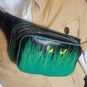 May include: A black leather fanny pack with a green and yellow grass design. The pack has a zipper closure and white stitching.