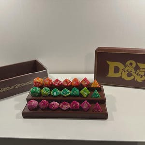 May include: A selection of multi-sided dice in various colours, arranged on a tiered wooden stand. A brown box with a gold D&D logo and a matching wooden box with decorative trim are also present.