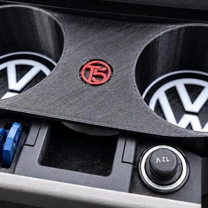 May include: A black 3D-printed dual cup holder insert for a vehicle dashboard, featuring two circular openings with white Volkswagen logos at the base and a red T5 emblem in the centre. The unit is positioned above a 12V power outlet and a small storage tray.