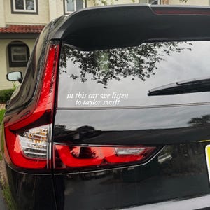 Könnte beinhalten: Ein weißer Vinyl-Aufkleber auf der Heckscheibe eines schwarzen Fahrzeugs. Der Aufkleber zeigt den Text in this car we listen to taylor swift in einer kursiven Schriftart. Er ist auf der Scheibe oberhalb der roten Rückleuchte angebracht.