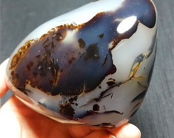 May include: A polished, oval-shaped stone with a smooth surface. The stone features a blend of white, dark brown, and deep blue colors. The colors are swirled and layered, creating a unique pattern. The stone is held in a hand.