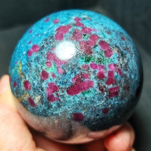 Puede incluir: Una piedra esférica pulida que presenta un color base azul turquesa vibrante con inclusiones cristalinas de color magenta intenso y rojo, acentuada por pequeñas manchas de depósitos minerales de color verde oscuro en la superficie lisa.