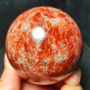 May include: A polished, spherical sunstone featuring a vibrant, mottled pattern of deep orange, reddish-brown, and creamy white mineral inclusions. The surface exhibits a smooth, reflective texture with a bright, centralized light reflection.