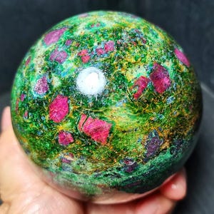 May include: A polished spherical mineral specimen featuring a vibrant green matrix with embedded deep pink and red crystalline inclusions. The surface displays a smooth, glossy texture with natural color variations and a bright light reflection.