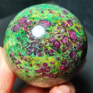 Op de afbeelding: Een gepolijste bolvormige steen met een levendige mix van diep magenta-rode kristalinsluitingen ingebed in een heldergroene matrix. Het oppervlak vertoont een natuurlijke, gevlekte textuur met verspreide goudgele minerale aders.