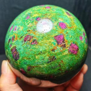 以下が含まれることがあります： 鮮やかな緑色のベースに、濃いマゼンタ色と赤色の結晶インクルージョンが埋め込まれた球状の鉱物標本。表面は質感のあるまだら模様で、全体に細く不規則な金褐色の筋が入っています。