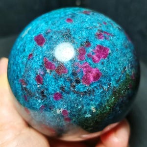 Peut inclure: Une pierre précieuse sphérique polie présentant une couleur de base bleu turquoise vibrante avec des inclusions cristallines rose foncé et magenta, accentuée par des taches minérales sombres et une surface lisse et réfléchissante.