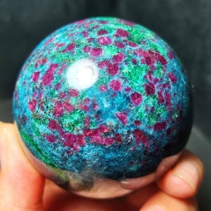 Puede incluir: Una esfera mineral pulida que presenta una mezcla vibrante de inclusiones de color rojo magenta intenso incrustadas en una matriz de material cristalino de color verde esmeralda y azul cerceta brillante, con una superficie lisa y reflectante.