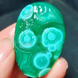 May include: An oval-shaped polished malachite gemstone featuring a vibrant green base colour with distinct, concentric circular patterns in lighter shades of teal and cyan throughout the surface of the stone, showcasing natural banding.