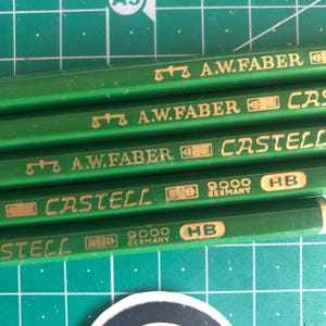 May include: Close-up of several green pencils with gold lettering. The pencils are labeled "A.W.FABER" and "CASTELL 9000 HB GERMANY". The pencils are arranged on a green cutting mat.