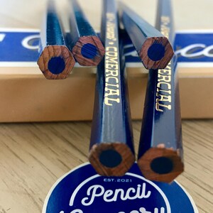 Puede incluir: Primer plano de cinco l&aacute;pices hexagonales azules con letras doradas que dicen "COMMERCIAL" y "EST. 2021". Los l&aacute;pices est&aacute;n afilados y tienen una mina azul oscura expuesta.