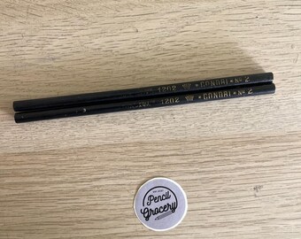 Puede incluir: Dos lápices de madera negros con letras doradas que dicen 1202, un icono de corona y CONDRI No 2. Debajo de los lápices hay una pegatina circular con un fondo gris claro y texto oscuro que dice est. 2021 Pencil Grocery con un icono de lápiz.