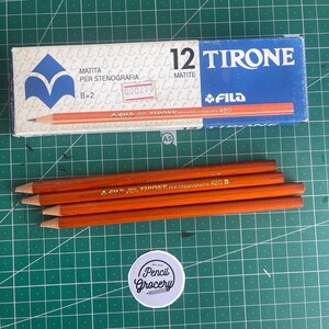 Peut inclure: Une boîte de 12 crayons de sténographie Fila Tirone avec un emballage bleu et blanc portant le texte 12 MATITE TIRONE et quatre crayons en bois orange avec une inscription dorée Fila Tirone per stenografia 420 B, disposés horizontalement.