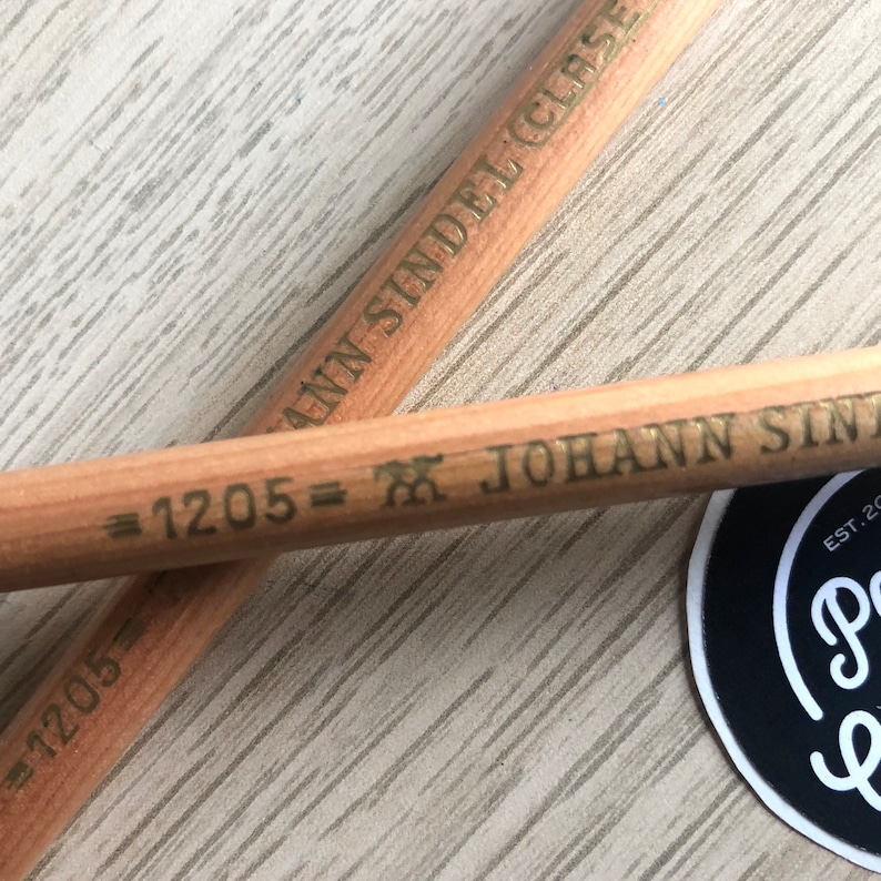 K&ouml;nnte beinhalten: Zwei Bleistifte aus Holz mit goldener Beschriftung. Die Stifte sind mit "=1205=XJOHANN SINDEL (CLASE" und "=1205=XJOHANN SIN" beschriftet.