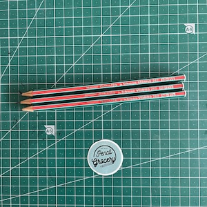 May include: Three green and red pencils with the text Schwan-STABILO 282 HB=2 1/2 Othello Germany printed on the side. Below the pencils is a circular sticker with a light blue background and the black text Pencil Grocery with a pencil icon.