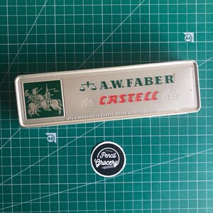 May include: A rectangular silver pencil case with the text "A.W. FABER CASTELL" in green and red. The case has a green square with a knight on a horse. A round sticker with the text "Pencil Grocery" is below the case.
