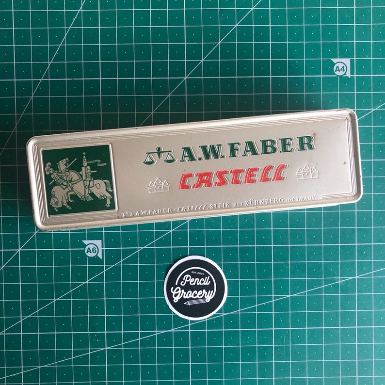 May include: A rectangular silver pencil case with the text "A.W. FABER CASTELL" in green and red. The case has a green square with a knight on a horse. A round sticker with the text "Pencil Grocery" is below the case.
