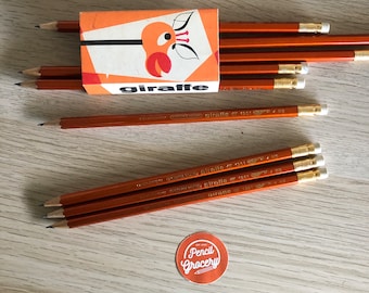 Puede incluir: Una caja de lápices naranjas con la palabra "jirafa" impresa en la parte delantera. Los lápices son naranjas con detalles dorados y tienen un diseño de jirafa en la goma de borrar.