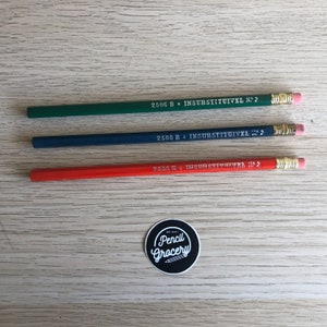 Op de afbeelding: Drie potloden, een groene, een blauwe en een rode, met de tekst "2506 B INSUBSTITUIVEL N° 2" erop gedrukt. Een zwart-witte sticker met de tekst "Pencil Grocery" bevindt zich in het onderste midden van de afbeelding.