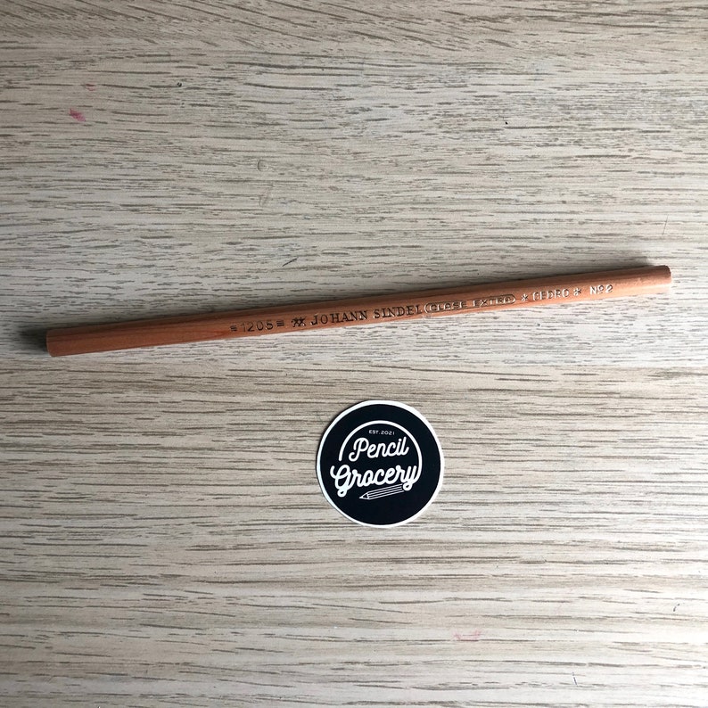 K&ouml;nnte beinhalten: Ein brauner Holzstift mit dem Aufdruck "#1205 = M. JOHANN SINDELGRABEN 131/D  +  G.D.R.O.C.  N92". Der Stift liegt auf einer hellbraunen Holzoberfl&auml;che. Ein schwarz-wei&szlig;er Aufkleber mit dem Text "Pencil Grocery" befindet sich in der unteren rechten Ecke des Bildes.