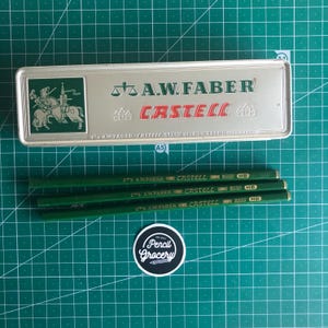 May include: A silver pencil case with the text "A.W. Faber Castell" and a knight illustration, along with several green pencils. The pencils are labeled "A.W. Faber Castell HB". A "Pencil Grocery" sticker is also visible.