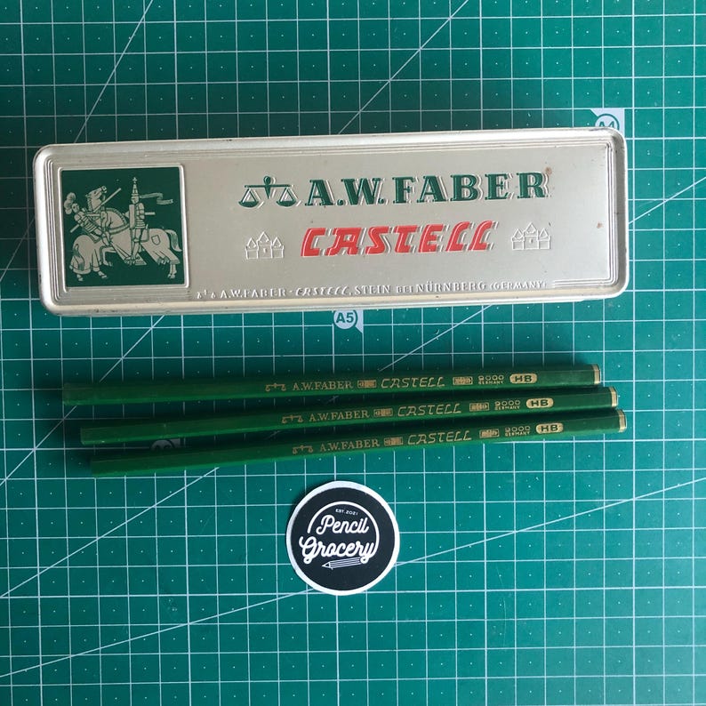 May include: A silver pencil case with the text "A.W. Faber Castell" and a knight illustration, along with several green pencils. The pencils are labeled "A.W. Faber Castell HB". A "Pencil Grocery" sticker is also visible.