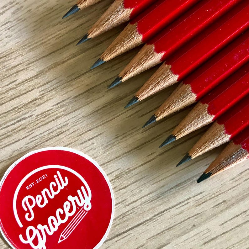Może przedstawiać: Zbliżenie na grupę czerwonych oł&oacute;wk&oacute;w z czarnymi końc&oacute;wkami grafitowymi. Oł&oacute;wki są ułożone wachlarzowo na drewnianej powierzchni. Czerwona okrągła naklejka z tekstem "Pencil Grocery" i "Est. 2021" znajduje się w lewym dolnym rogu obrazu.