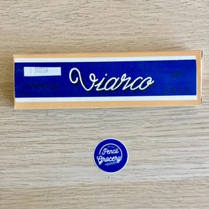 Puede incluir: Una caja azul y amarilla con el texto "1 DUZIA COMMERCIAL" y "Viarco" en cursiva. La caja est&aacute; sobre una superficie de madera.