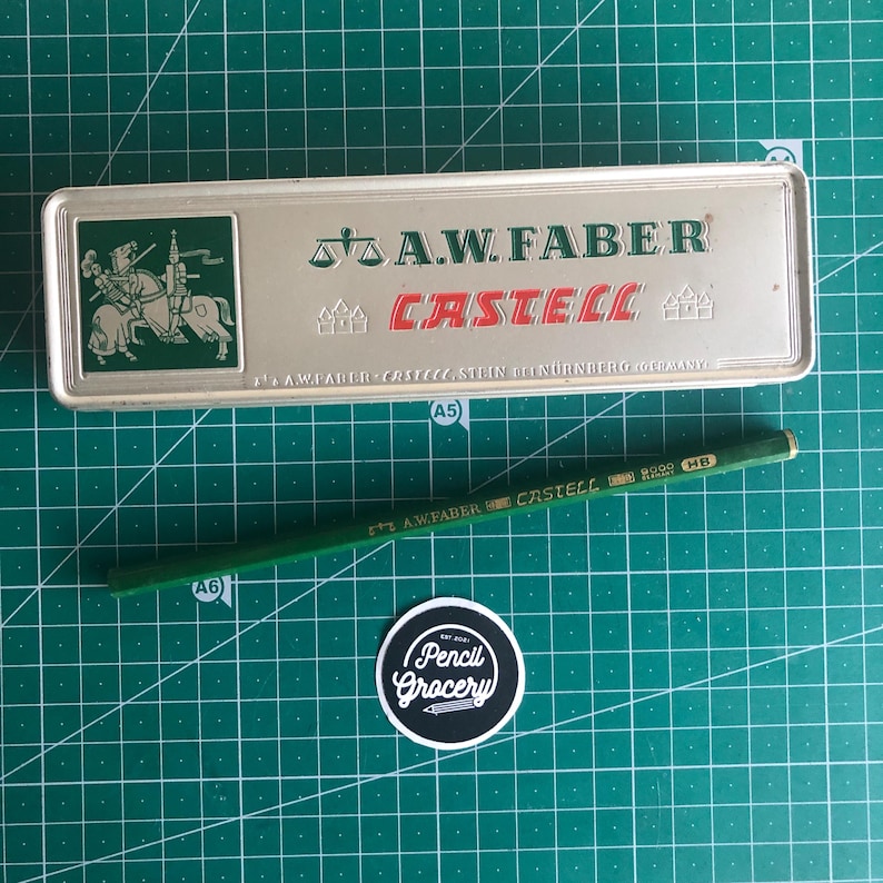 May include: A silver pencil case with the text "A.W. FABER CASTELL" and a green pencil. The pencil case has a green emblem of a knight on a horse. The pencil is green with the text "A.W. FABER CASTELL HB". A sticker with the text "Pencil Grocery" is also visible.