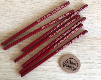 Puede incluir: Varios lápices rojos con la inscripción "250 * Marco" y "DESENHO N:2". También hay un disco de madera con la inscripción "Pencil Grocery". Los lápices están dispuestos en diagonal.