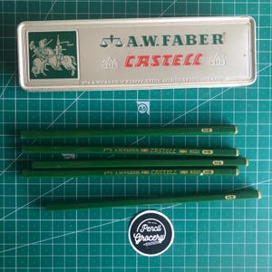 May include: A set of green pencils with gold tips and the text "HB" are arranged near a metal pencil case. The case is silver with the text "A.W. FABER CASTELL" and a knight illustration. A "Pencil Grocery" sticker is in the foreground.