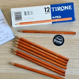 Può includere: Un assortimento di matite arancioni con punte in grafite nera, insieme a una scatola blu e bianca con la scritta "12 TIRONE MATITE" e un adesivo "Pencil Grocery". Le matite sono contrassegnate "FILA TIRONE PER STENOGRAFIA 420 2B".