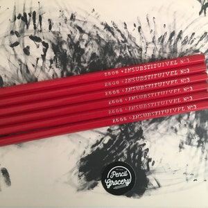 Puede incluir: Seis lápices rojos con letras blancas que dicen "2506 INSUBSTITUIVEL N:3" en cada lápiz. Los lápices están apilados en una superficie blanca con huellas de manos negras.