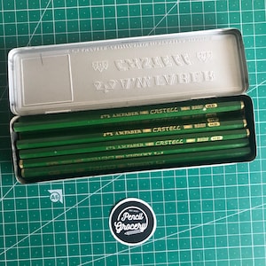 May include: A metal pencil case containing several green pencils. The pencils are labeled "A.W. Faber Castell 8900 HB". The case is open, revealing the pencils inside. The background is a green cutting mat.