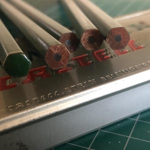 May include: Close-up of several hexagonal pencils with a green end and a dark gray body. The pencil tips are visible, showing the graphite core. The pencils are resting on a metal case with red lettering.