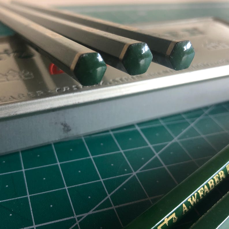 May include: Close-up of three gray pencils with green hexagonal tips. The pencils are resting on a metal case and a green cutting mat. The pencils have a silver band near the green tip.