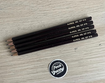 Puede incluir: Cinco lápices de madera de color marrón oscuro con puntas afiladas, cada uno con el texto impreso en oro FILA 117 N°3 MADE IN ITALY. Debajo de los lápices hay una pegatina circular negra con borde blanco y el texto blanco Pencil Grocery EST. 2021.