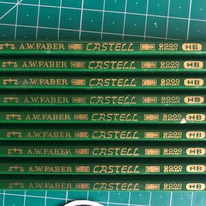 May include: A close-up shot of several green pencils with gold lettering. The pencils are labeled "A.W. Faber Castell 9000 HB Germany". The pencils are arranged in parallel rows on a green surface.